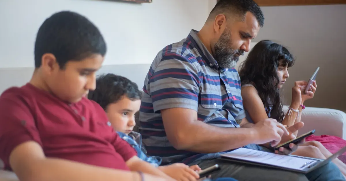 Um pai e três filhos no sofá, cada um usando celular ou tablet