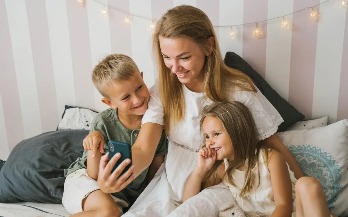 Maminka se dvěma dětmi se na posteli společně dívají do mobilu